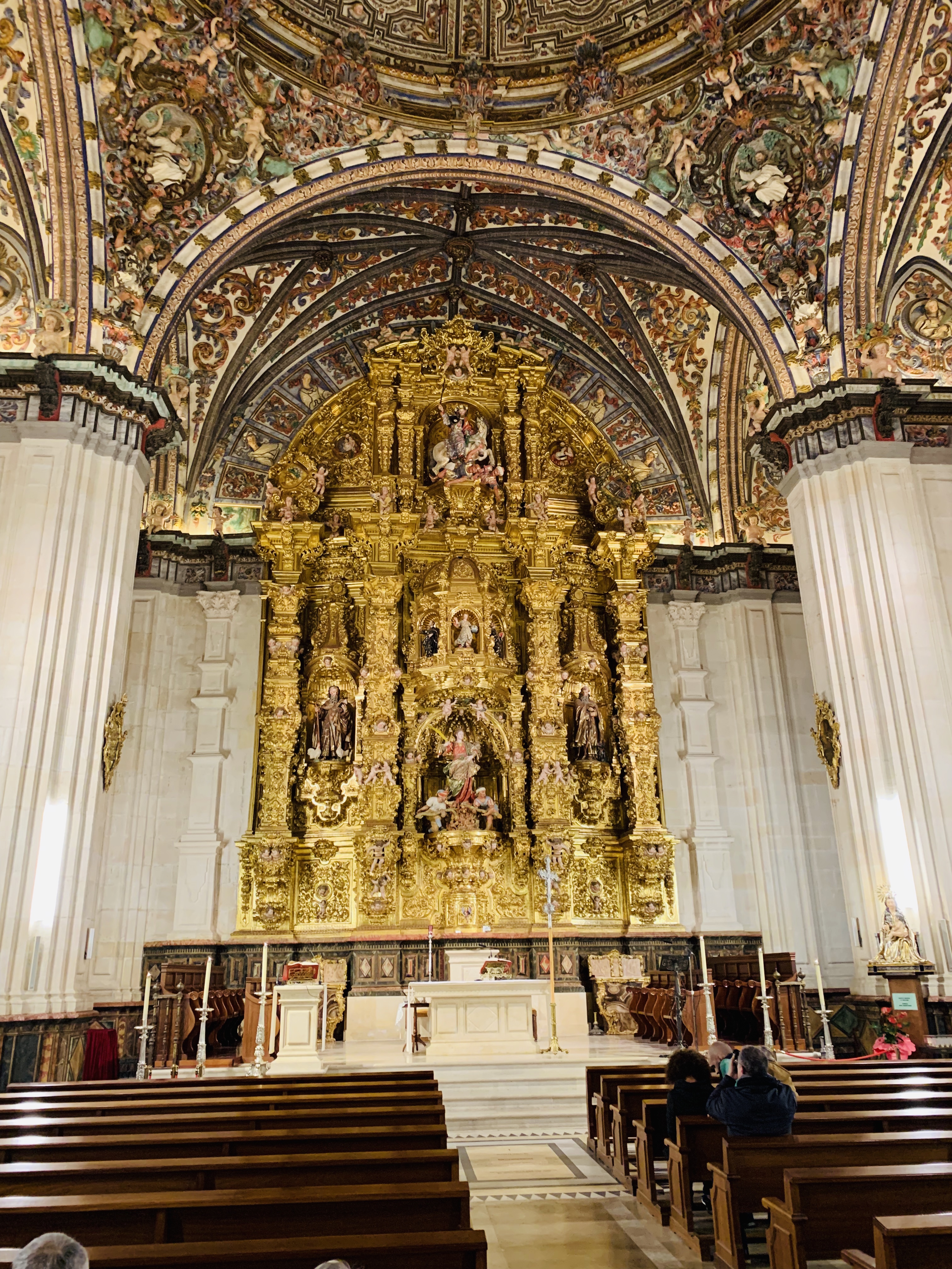 Interior of a richly decorated chapel featuring a gilded altar, intricate ceiling frescoes, and wooden pews.