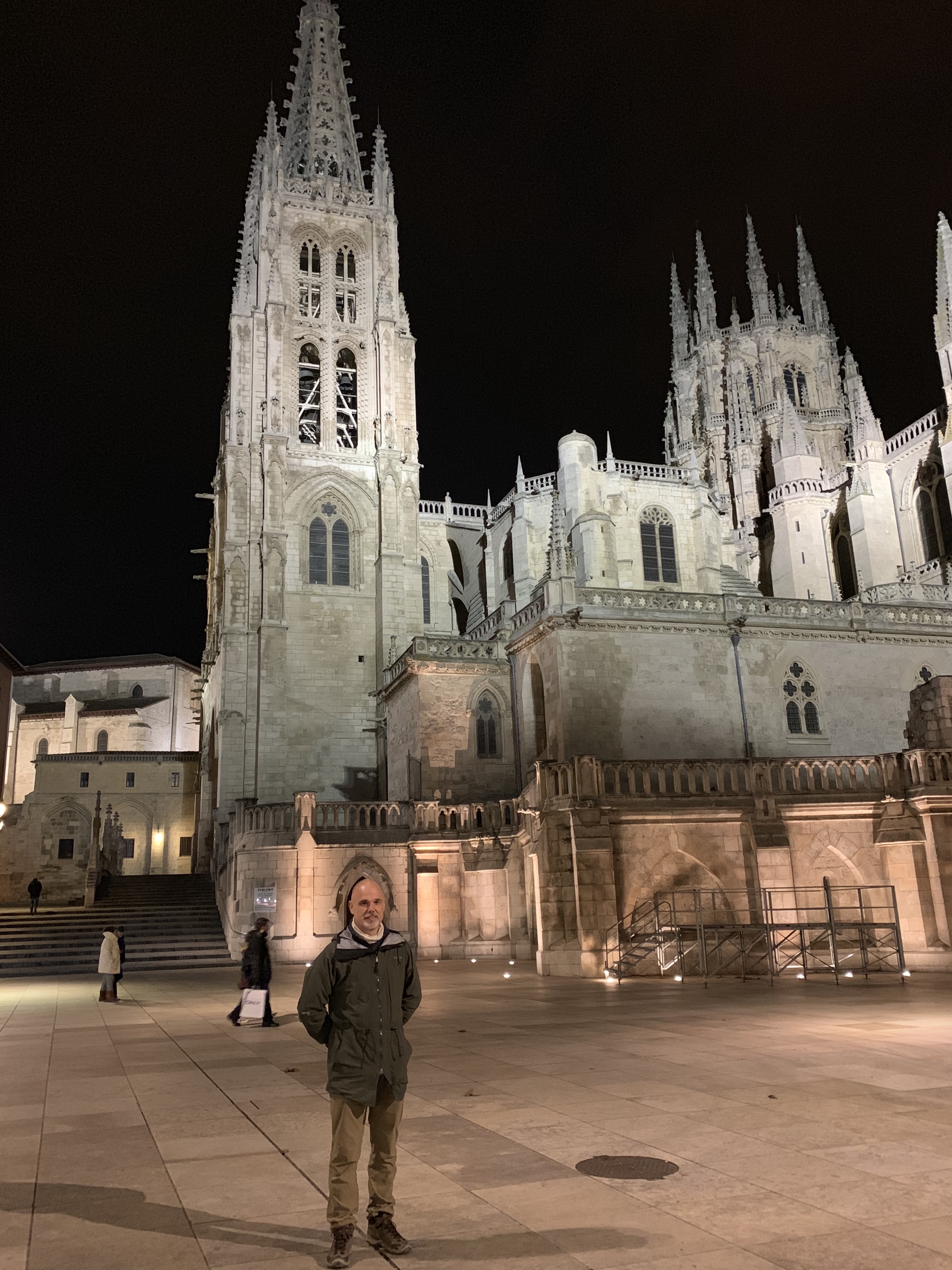 A man standing in front of the Burgos Cathedral at night, showcasing its illuminated Gothic architecture.