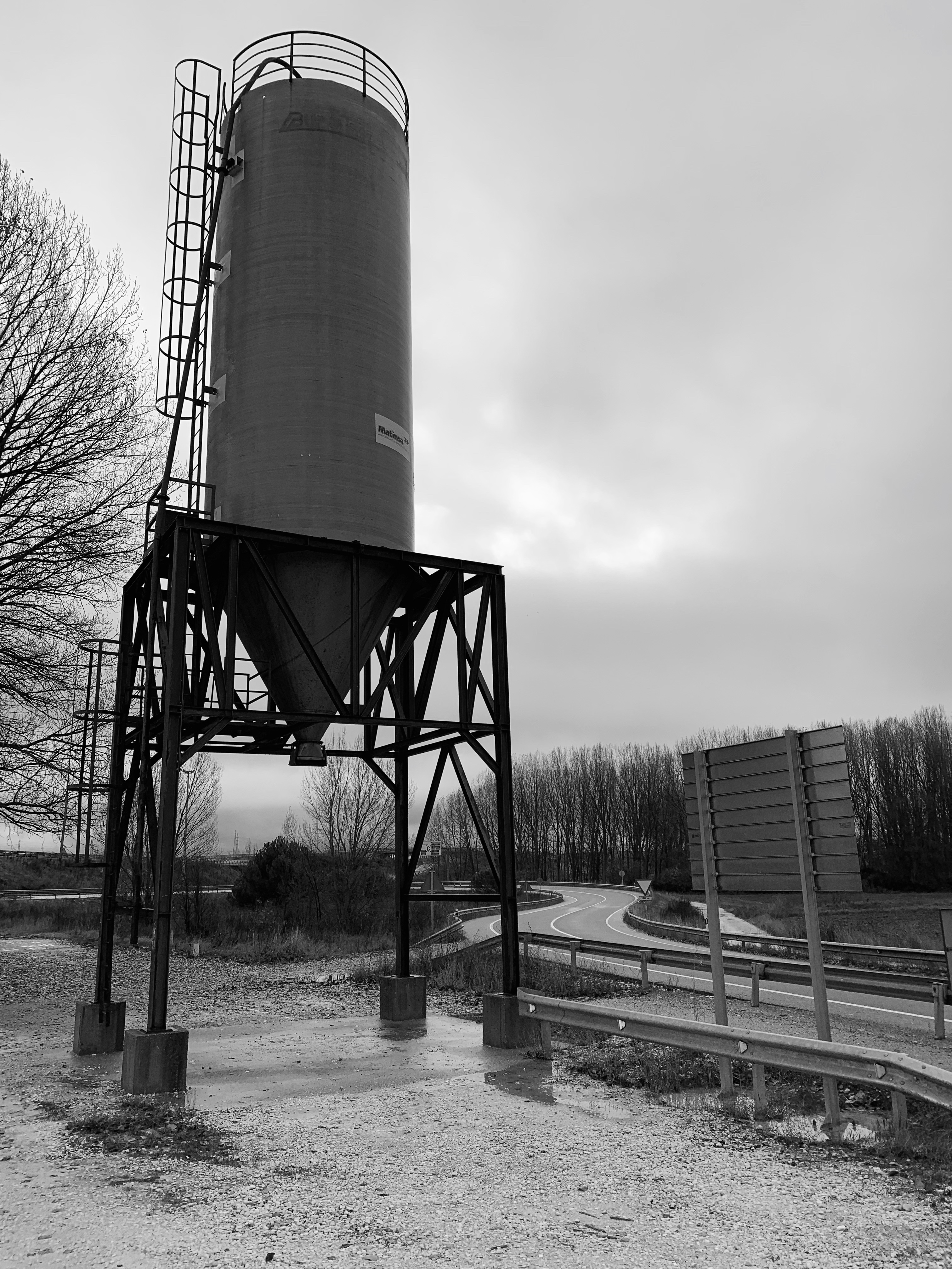 A tall industrial silo on a metal frame beside a winding road, captured in black and white with overcast skies in the background.