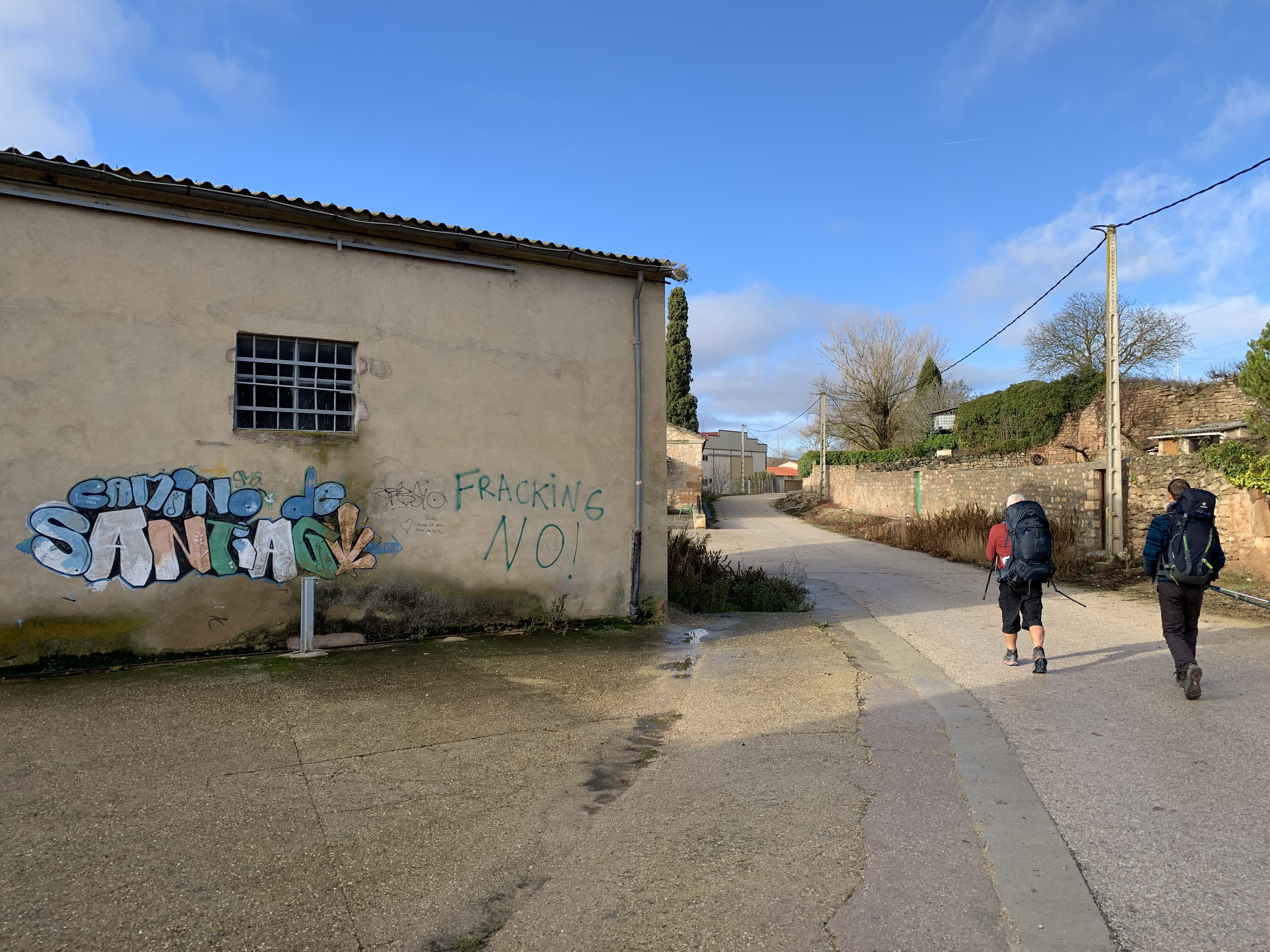 Pilgrims walking along a road near a wall with graffiti that reads 'camino de Santiago' and 'FRACKING NO!' in bold letters, under a partly cloudy blue sky.
