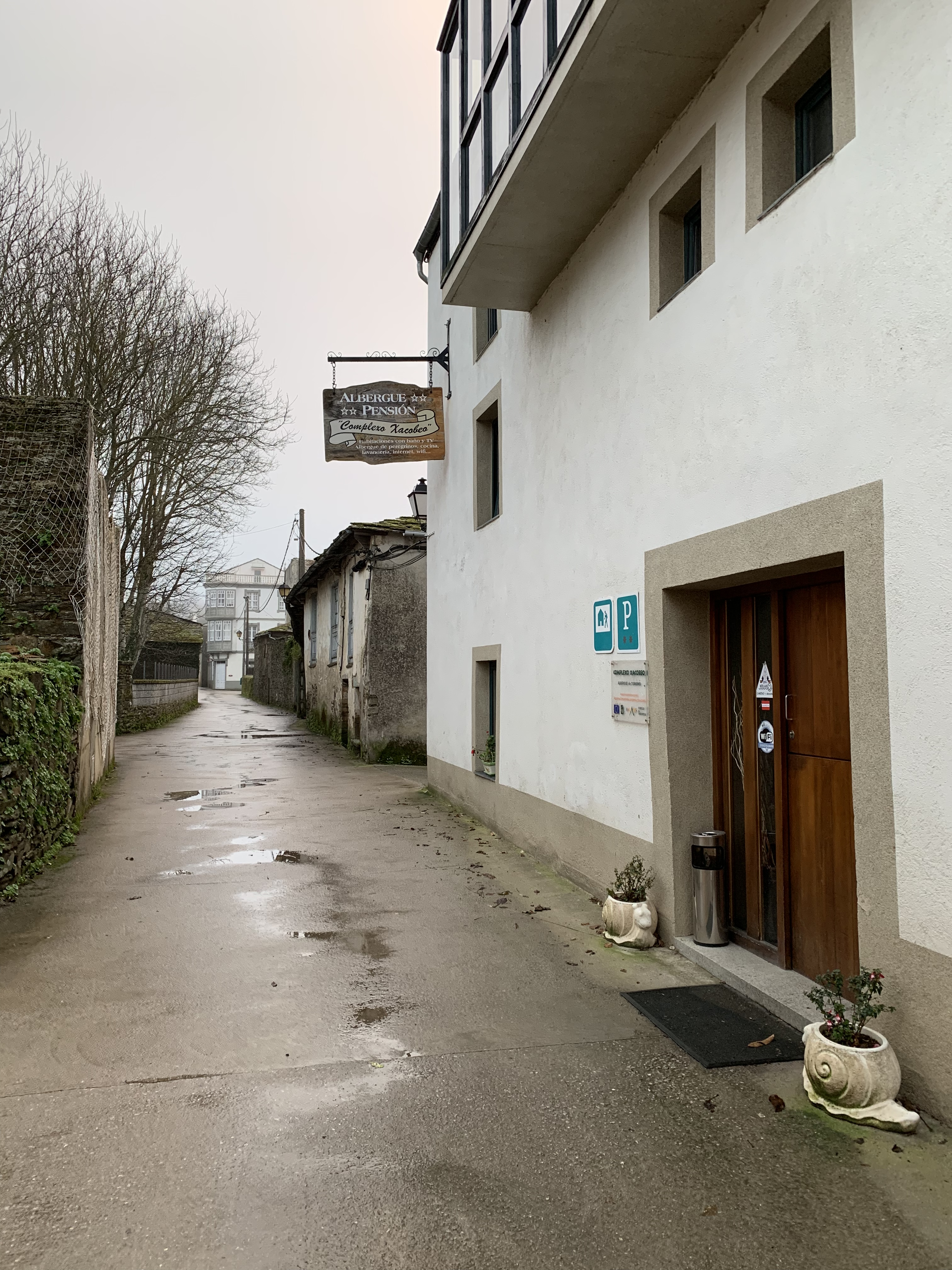 A narrow street in a quaint village with a sign for an albergue and pension, showcasing a rustic building with a wooden door and planters at the entrance.