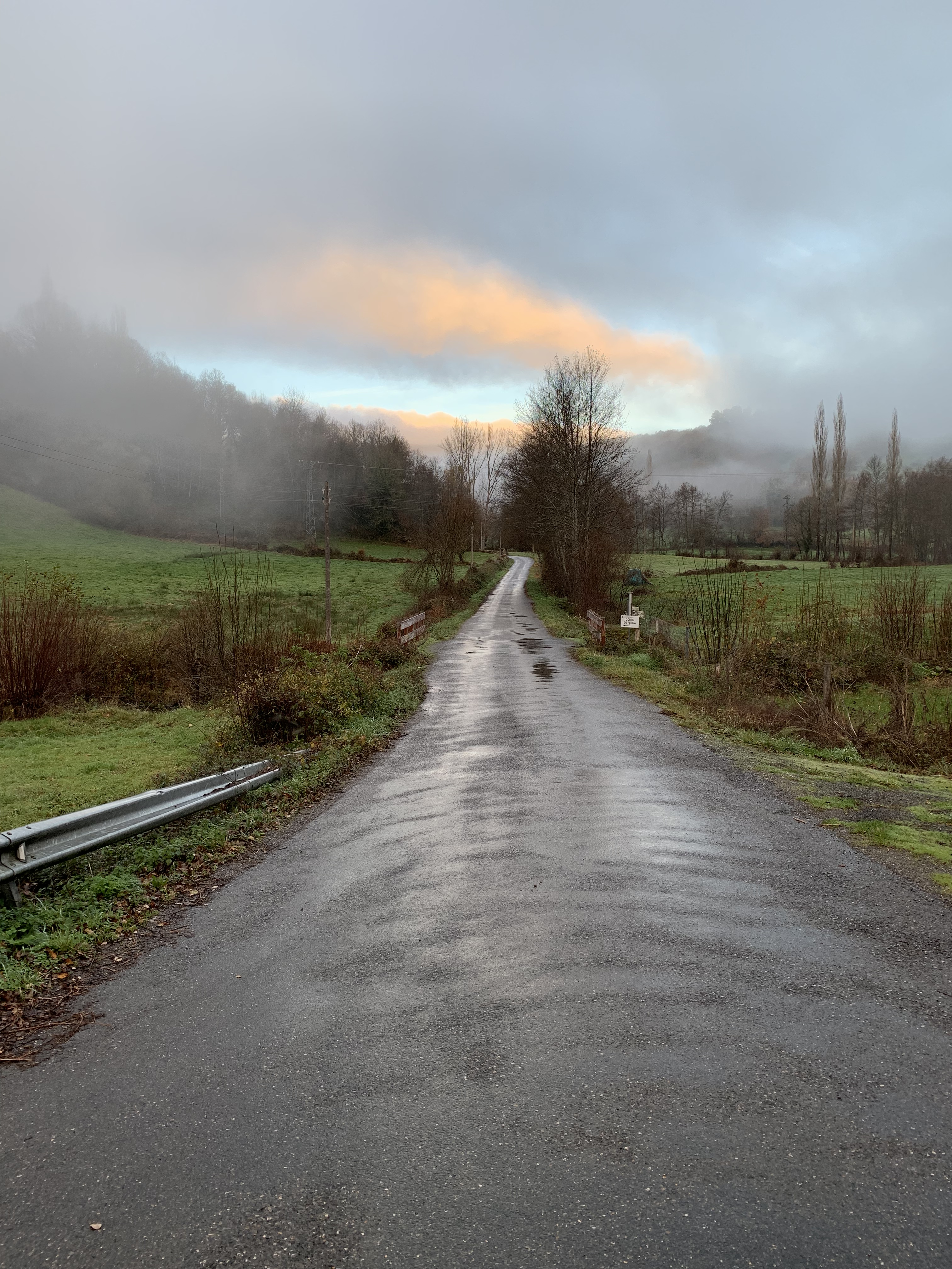 A winding road through a misty landscape, with fog covering the hills and a hint of orange in the sky as the sun rises.