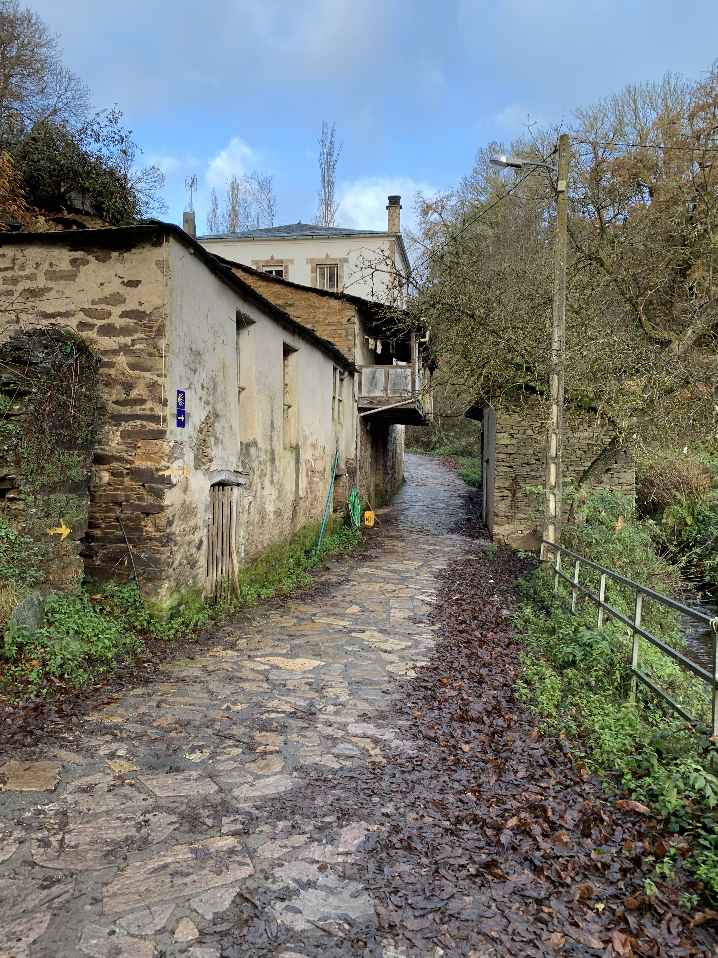 A cobblestone pathway winding past rustic stone buildings, with patches of greenery and a clear blue sky above.