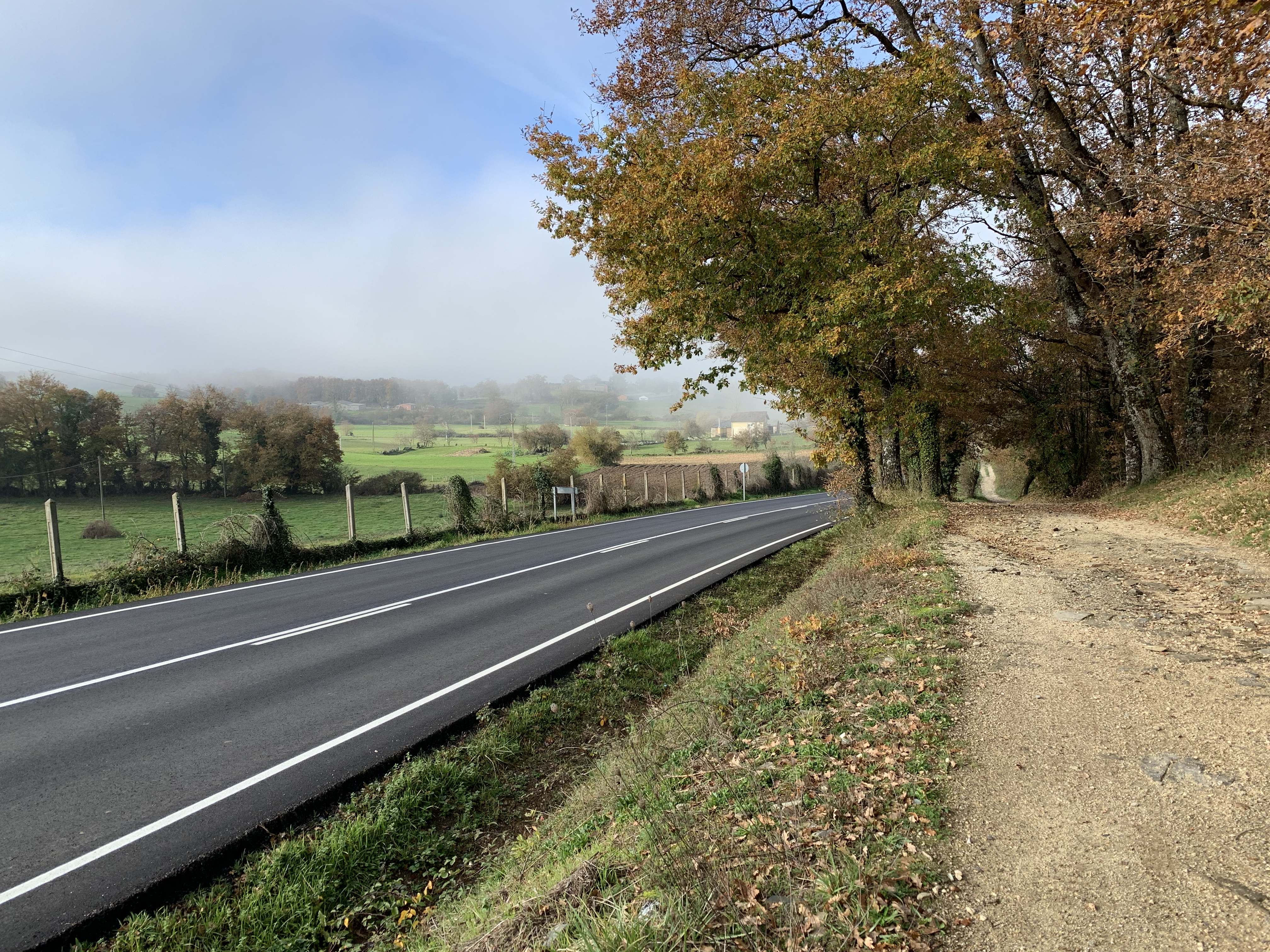 A scenic view of a countryside road alongside a dirt path, bordered by trees with autumn foliage and rolling green fields in the background, partially shrouded in mist.